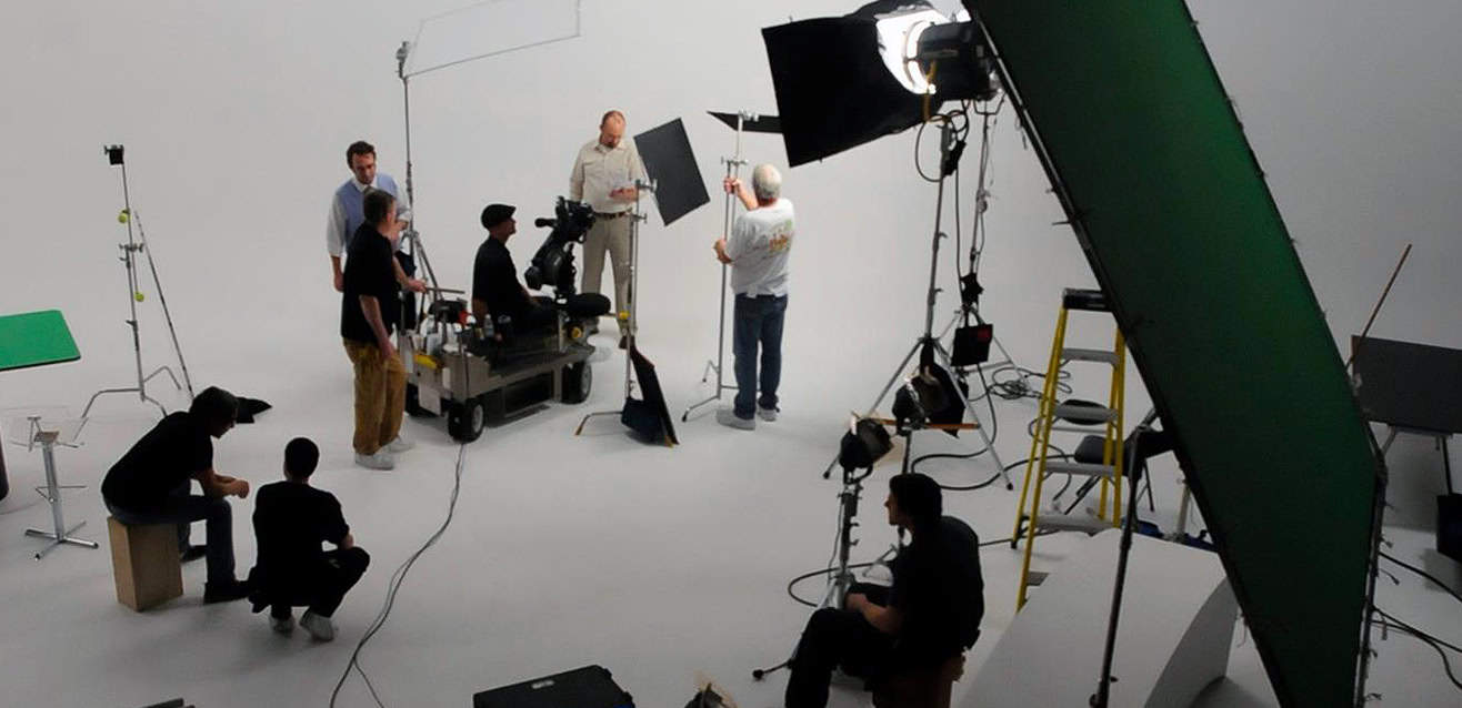 Behind-the-scenes view of a studio production set with crew members, professional lighting, a green screen, and a camera operator on a dolly preparing for a shoot.