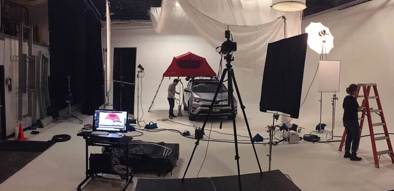 Studio setup featuring a car with a rooftop tent, surrounded by lighting equipment, tripods, and a crew member adjusting a ladder, with a computer monitor displaying the live shot.