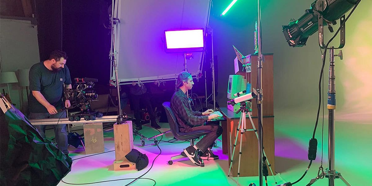 Behind-the-scenes studio setup with a videographer operating a camera on a dolly track and a subject seated at a desk under vibrant, colored lighting, showcasing the versatility of professional camcorders like the Sony PMW-EX3 for creative productions