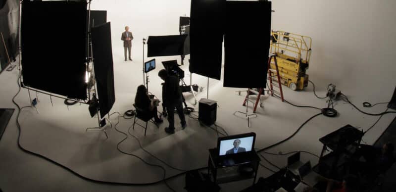 Professional studio setup with lighting equipment, a camera operator, and a presenter standing in front of a white backdrop during video production.