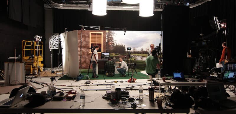 Behind-the-scenes of a video production set with a studio backdrop, multiple cameras, crew members working, lighting equipment, and a table filled with laptops, cables, and production tools.