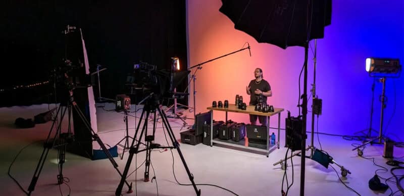 Video production studio with a presenter at a table showcasing camera lenses, surrounded by professional lighting, tripods, and a boom microphone against a colorful gradient backdrop.