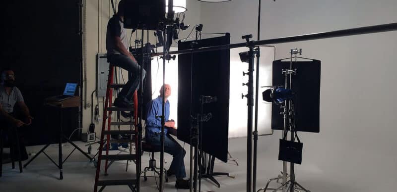 A professional video production setup with lighting, diffusers, and crew working around a seated subject in a studio environment.