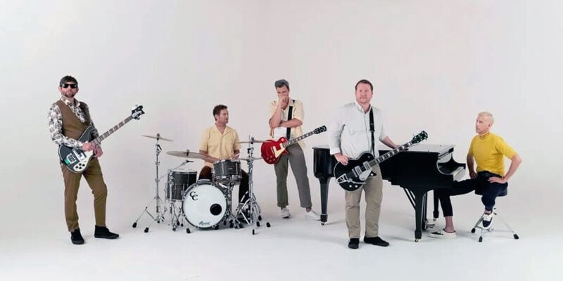 Band posing with guitars, drums, and a piano in a minimal sound stage setup at Pixthis in Portland, showcasing a clean and versatile filming environment with a white cyclorama background.