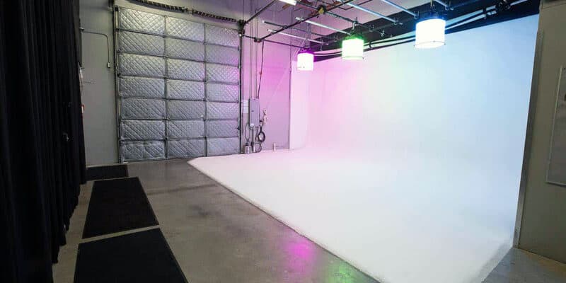 Empty sound stage at Pixthis in Portland, featuring a white cyclorama wall, large roll-up door, overhead lighting grid, and black curtains, ready for professional video production setups.